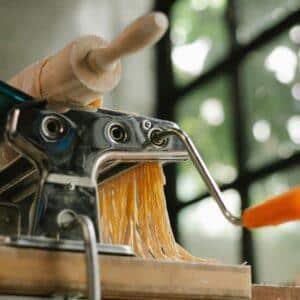 From below of modern stainless pasta maker equipment with cut raw dough placed on wooden table in kitchen on blurred background